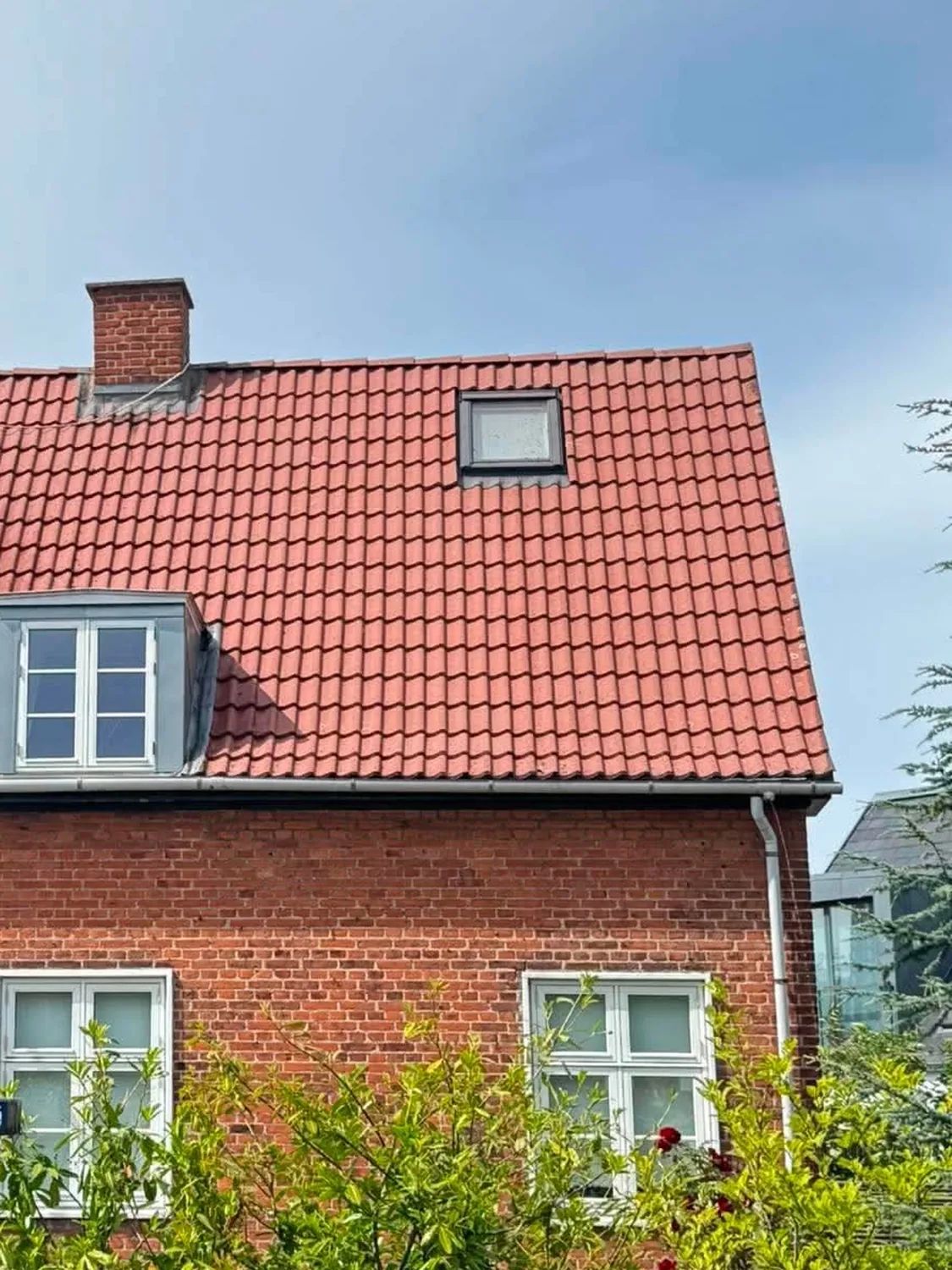 Rød murstensvilla med kvistvindue og ensartet rødt tegltag efter tagrens, fotograferet fra haven under en let skyet himmel